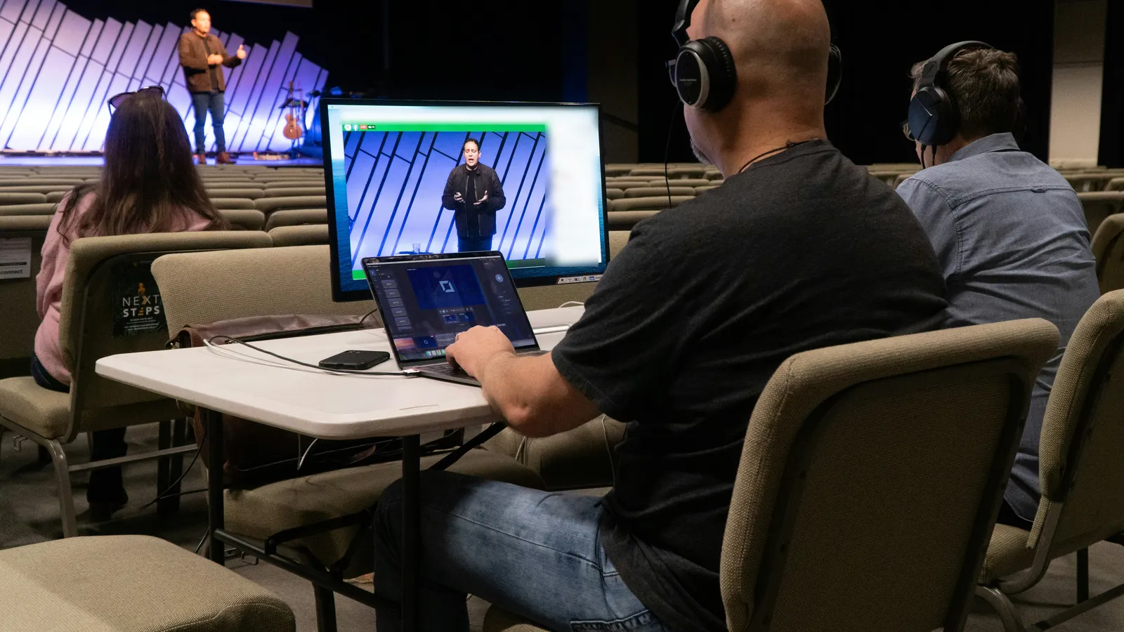 Two volunteers in headphones run the livestream and Verse Copilot from a table in the sanctuary while the pastor preaches